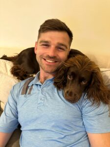 A man looks toward the camera smiling, a brown dog is draped across his shoulders.