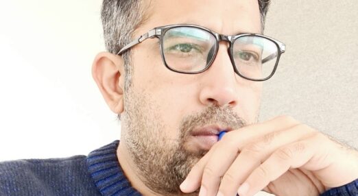 A man wearing glasses is holding a pen to his lips.