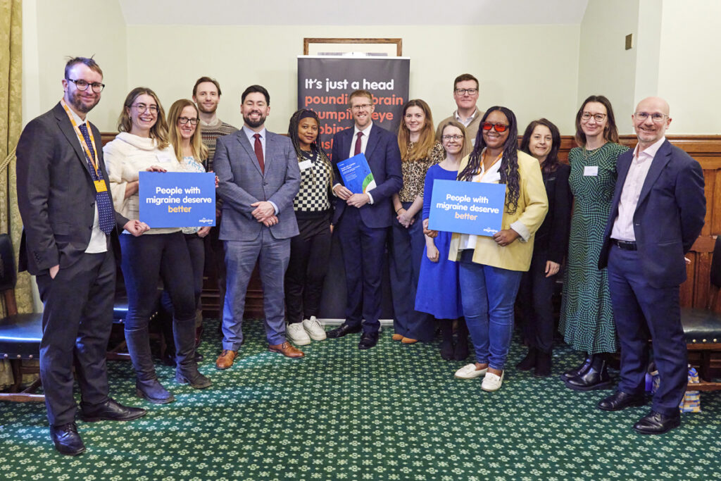 A group of MPs and patient advocates posing for a photo in the House of Commons. Two are holding signs that say people with migraine deserve better