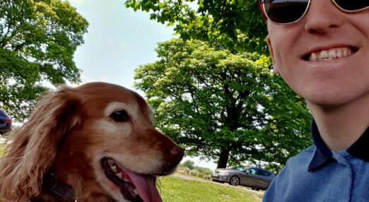 A man in sunglasses sitting on grass smiles alongside a golder retriever dog