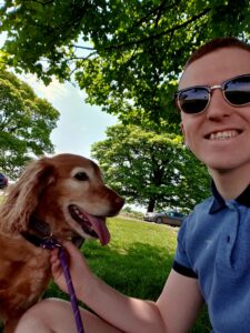 A man with sunglasses sitting on the grass smiles alongside a golden retriever dog 