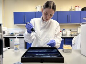 Mignon is in a laboratory using a long pipette in front of a case full of slides 