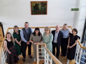 A group of 8 people stand on some stairs smiling at the camera. They are employees at NHS Grampian and Rob Music from The Migraine Trust