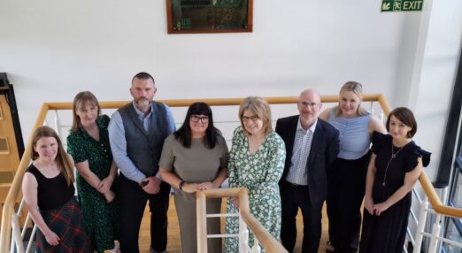 A group of 8 people stand on some stairs smiling at the camera. They are employees at NHS Grampian and Rob Music from The Migraine Trust