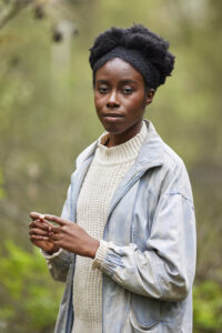 A woman wearing a white jumper and denim jacket with black hair tied up looks at camera