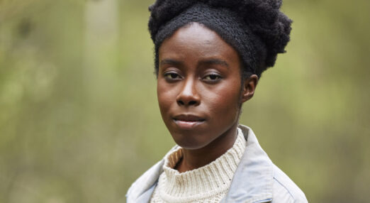 A woman wearing a white jumper and denim jacket with black hair tied up looks at camera