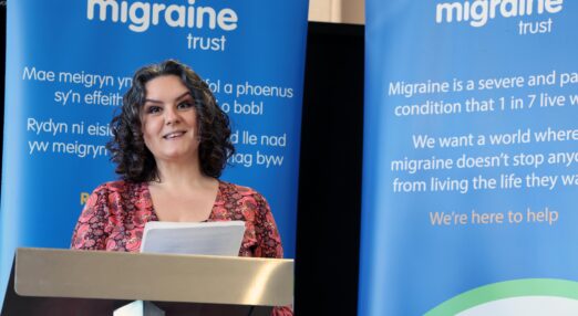 A woman with migraine is speaking at an event at a lectern in front of 2 blue banners promoting The Migraine Trust