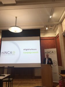 A man in a suit stands behind a podium, in front of a presentation screen that reads NINCA #FightForNeuro