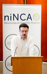 A man in a white shirt and glasses stands behind a podium, witha banner stand reading 'NINCA' in the background.