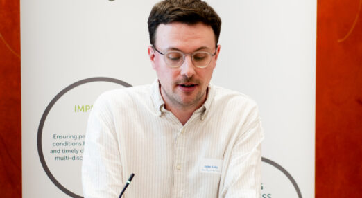 A man in a white shirt and glasses stands behind a podium, witha banner stand reading 'NINCA' in the background.