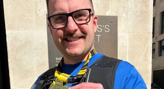 A man wearing glasses is smiling at the camera. He is wearing a medal and holding it in his hand.