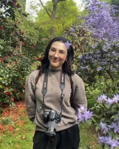 A young woman is wearing a camera around her neck and has a purple flower in her hair. She is smiling at the camera.