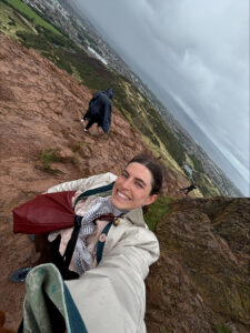 A woman stands on top of a hill, in the background is a scenic Scottish landscape
